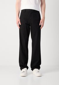 Black, loose-fitting trousers made from cotton, featuring a button closure, front pockets, and a smooth texture. Worn with white sneakers.