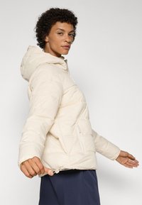 Veste matelassée légère avec capuche, dotée d'une texture piquée, de poches avant zippées et d'un ourlet ajustable avec cordon.