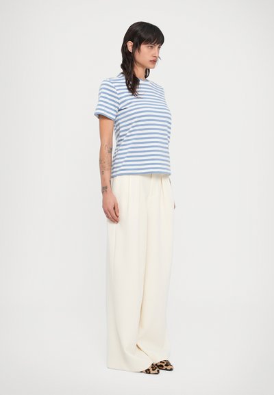 Γυναίκα που στέκεται φορώντας μπλε και λευκό ριγέ κοντομάνικο πουκάμισο, φαρδιές λευκές παντελόνες και μυτερά παπούτσια με λεοπάρ σχέδιο, κοιτάζοντας προς τα δεξιά.