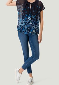 Chemisier noir à motifs floraux bleus et blancs, en tissu transparent, avec un col arrondi, associé à un jean bleu et des baskets en argent.