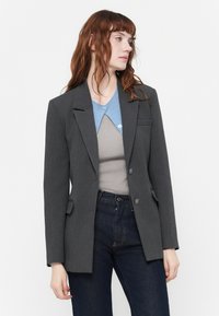 Blazer grigio su misura con revers a punta, due tasche frontali e chiusura a bottoni. Indossato sopra una camicia col celeste e jeans scuri.