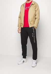 Beige Jacke mit Reißverschluss, rotes T-Shirt mit Grafik, schwarze Jogginghosen mit weißem Markenlogo und weiße Sneakers mit schwarzen Akzenten. Moderner Freizeitlook.