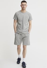 Grå bomulds t-shirt med kort ærme og brystlomme, parret med grå shorts. Modellen har blå sportsko og hvide sokker på.