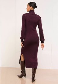 Tumši sarkana adīta kleita ar augstu apkakli, pinuma raksta garām piedurknēm un sānu šķēlumu. Valkāta ar brūnām ādas zābaciņām līdz ceļiem.