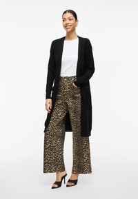 Svart lång kofta över en vit skjorta, kombinerad med vida leopardmönstrade byxor. Har öppen front, ribbade detaljer och högklackade muletofflor.