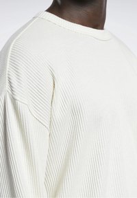 Zbliżenie na osobę w białym, teksturowanym swetrze z dzianiny, z okrągłym dekoltem i opadającym szwem ramienia.