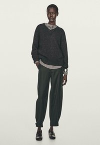 Dunkelgrauer Wollpullover mit V-Ausschnitt, über einem grauen Shirt getragen, kombiniert mit lockeren grünen Hosen und schwarzen Slippern. Glatter, strukturierter Stoff.