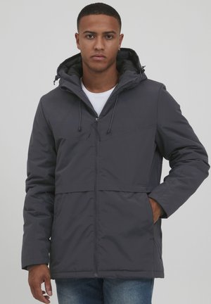 Veste à capuche grise avec fermeture éclair, deux poches avant et cordons de serrage ajustables. Portée par-dessus une chemise blanche. Texture de tissu lisse.