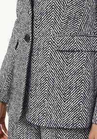 Schwarzes und weißes Holzfällerstoffmuster, das ein tailliertes Sakko mit einem einzelnen schwarzen Knopf und einer linken Brusttasche zeigt.