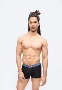 Άνδρας χωρίς πουκάμισο με μακριά dreadlocks που φοράει μαύρα μποξεράκια Tommy Hilfiger, στέκεται μπροστά σε απλό ανοιχτό γκρι φόντο.