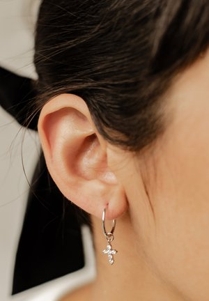 Silberne Creolen mit einem kleinen Kreuzanhänger, verziert mit klaren Steinen, die funkeln, getragen an einem Ohr, umrahmt von dunklem Haar, das das Gesicht umschmeichelt.