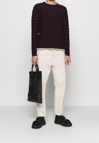 Personne portant un pull rayé noir et rouge, pantalon crème, sandales noires avec des chaussettes, tenant un sac fourre-tout en cuir noir à côté d'elle.