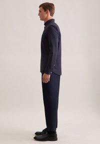 Uomo in camicia button-up blu navy e pantaloni dritti abbinati, in profilo contro uno sfondo chiaro e semplice.