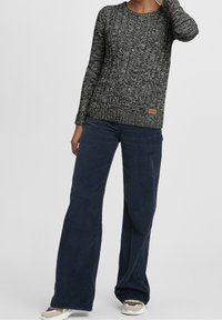 Pull en tricot à motifs noir et blanc avec des manches longues, associé à un pantalon large en velours côtelé bleu marine et des baskets beiges. Marquage sur le bas du pull.