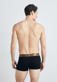 Black boxer briefs with a shiny gold waistband, smooth texture, and mid-thigh length, worn by a male model from the back.