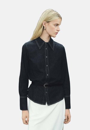 Camicia di denim scuro con maniche lunghe, colletto appuntito e cuciture bianche a contrasto, abbinata a una cintura nera in vita.