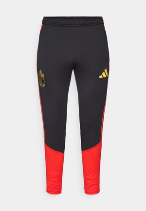 Čierne športové nohavice s červenými bočnými panelmi, s zlatým logom na ľavom stehne a žltým logom Adidas na pravej nohe.