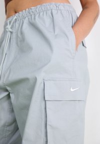 Light gray Nike cargo pants with elastic waistband, side pocket with flap, and a hand placed inside the pocket.