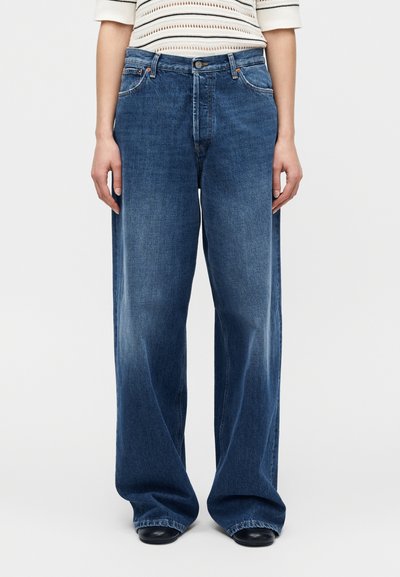 Femme portant un jean en denim bleu ample et un haut en maille blanc à manches courtes, debout devant un fond clair uni.