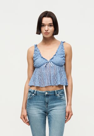 Joven con el pelo corto y oscuro, vestida con una camiseta corta de encaje azul y vaqueros azul claro, sobre un fondo liso.