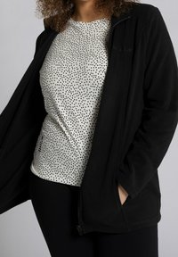 Giacca in pile nera con zip frontale, indossata sopra un top bianco con pois neri. Il modello indossa anche leggins scuri.