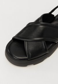 Mustat nahkakengät, joissa on ristikkäinen hihnasuunnittelu, pehmustettu pohja ja paksu kumipohja. Sileä pinta ja tikkaus yksityiskohtia.