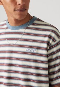 Prugasta majica s kratkim rukavima u krem, crvenim i plavim tonovima, s plavim rebrastim izrezom. Na prsima se nalazi ljubičasti Levi's logo. Izrađena od pamuka.