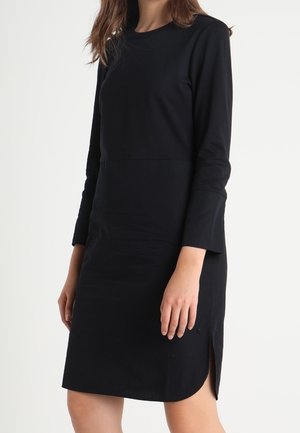 Frau trägt ein einfaches, schwarzes, knielanges Kleid mit langen Ärmeln und einem kleinen Seitenschlitz vor weißem Hintergrund.