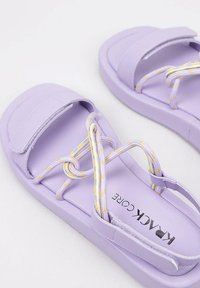 Sandalias moradas con un material sintético suave, correas de Velcro y un diseño de cuerda entrelazada en amarillo y blanco en la parte superior.