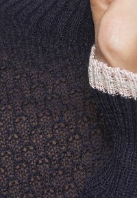 Die Hand ruht am Gesicht, während ein navyfarbener Strickpullover mit einem hellrosafarbenen Bündchen getragen wird.
