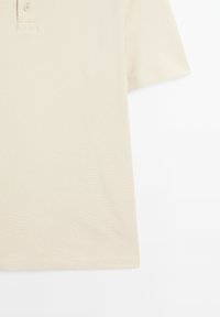 Beige polo tričko s krátkým rukávem, texturovaná tkanina, límec na dva knoflíky a rovný lem. Žádné vzory ani designy nejsou viditelné.