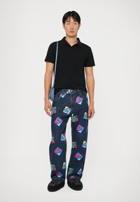 Cămașă polo neagră, blugi blue denim lejeri cu modele pătrate colorate în stil cartonat. Modelul poartă pantofi negri și o geantă de umăr bleu deschis.