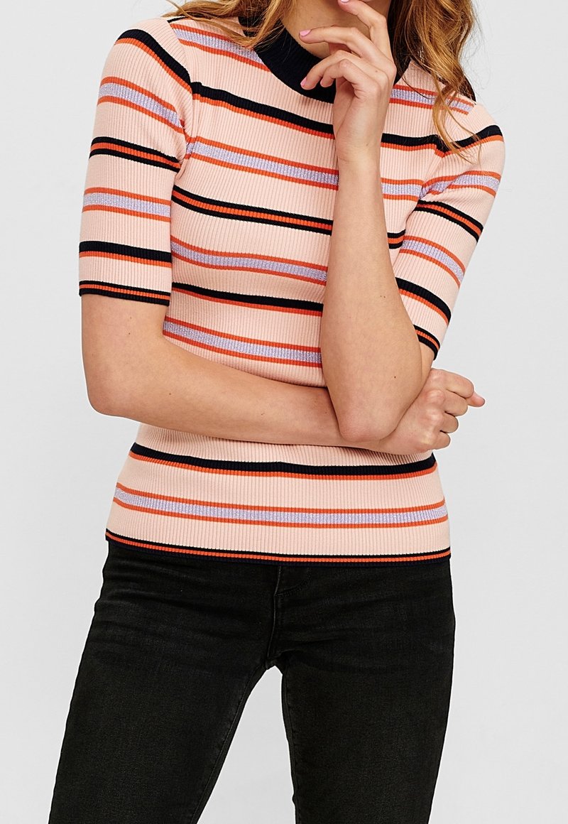 Femme portant un haut côtelé à manches courtes rayé en pêche, noir, orange et lavande, associé à un pantalon noir, bras croisés et main près du menton.