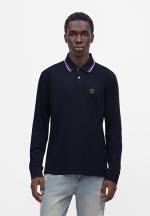 Jeune homme portant un polo bleu marine à manches longues avec un col rayé et un jean bleu clair déchiré sur fond blanc.