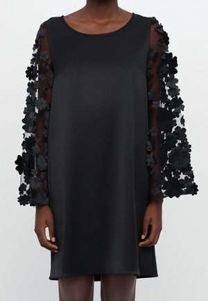 Czarna sukienka do kolan z przezroczystymi długimi rękawami ozdobionymi trójwymiarowymi aplikacjami kwiatowymi, noszona przez stojącą osobę z rozluźnionymi ramionami.