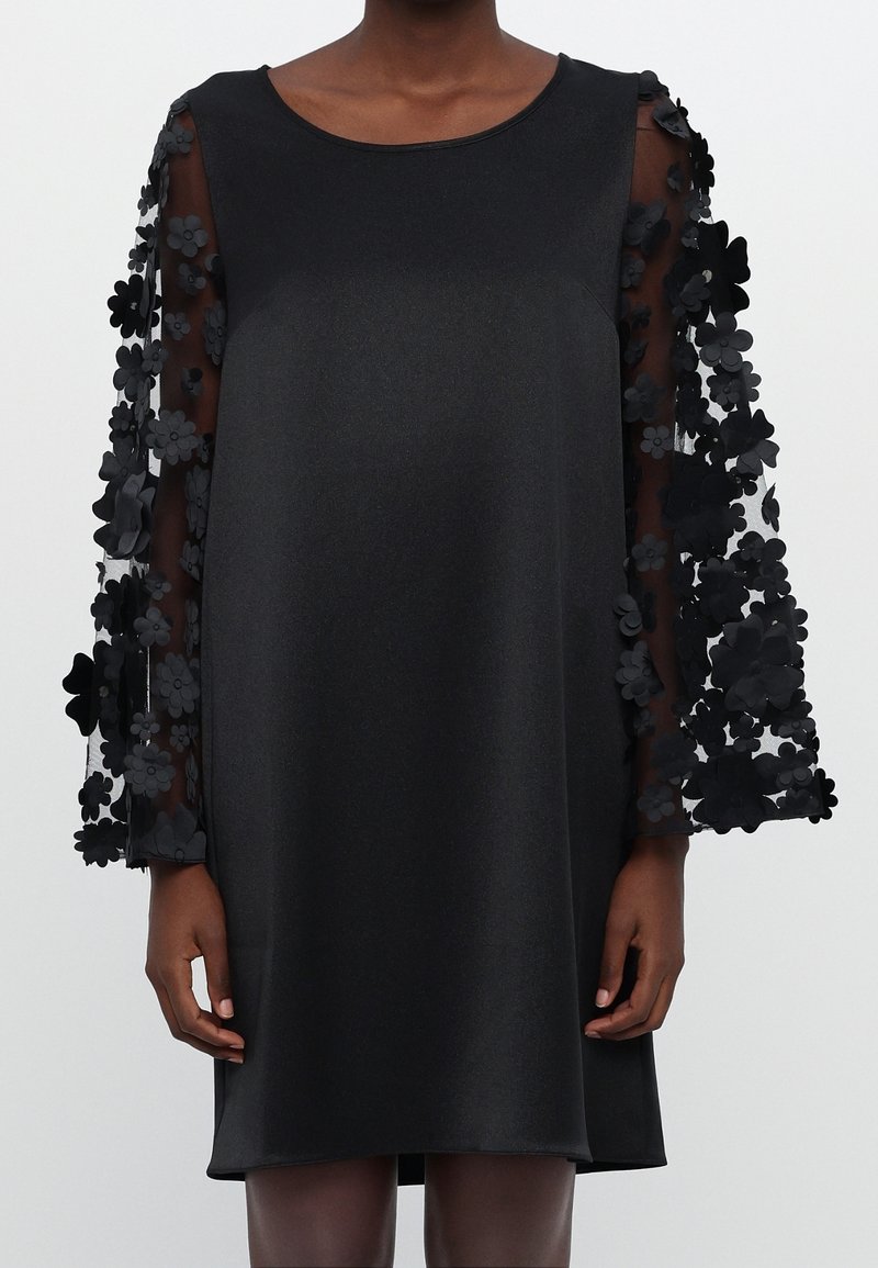 Robe noire au genou avec manches longues transparentes ornées d’applications florales 3D, portée par une personne debout avec les bras détendus.