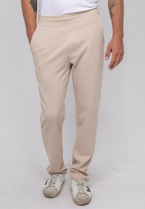 Homme portant un pantalon beige, une chemise blanche et des baskets blanches, debout les mains dans les poches sur un fond uni et clair.
