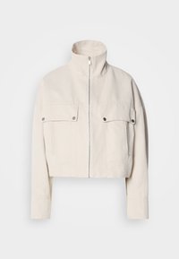 Κοντή μπεζ ζακέτα από βαμβάκι με ψηλό γιακά, κλείσιμο με φερμουάρ μπροστά και δύο τσέπες στο στήθος με κουμπιά ασφαλείας.