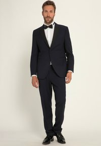 Tuxedo navy con revers in raso nero, camicia bianca e papillon nero. Vestibilità aderente, tessuto liscio, scarpe eleganti nere lucide.