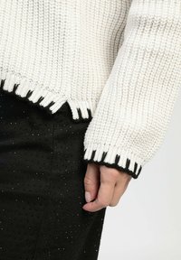 Pull en maille crème avec trame épaisse et bord noir, présentant une texture côtelée et un ourlet à franges, associé à un pantalon en tissu noir à petits pois.