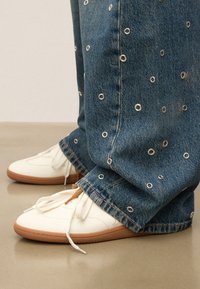 Baskets blanches avec une finition texturée et des accents marron, associées à un jean en denim bleu avec des œillets argentés et une coupe décontractée.
