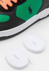 Baskets noires avec accents verts, matériau texturé, lacets roses et semelle en caoutchouc blanche. Comprend deux lacets blancs à côté de la chaussure.