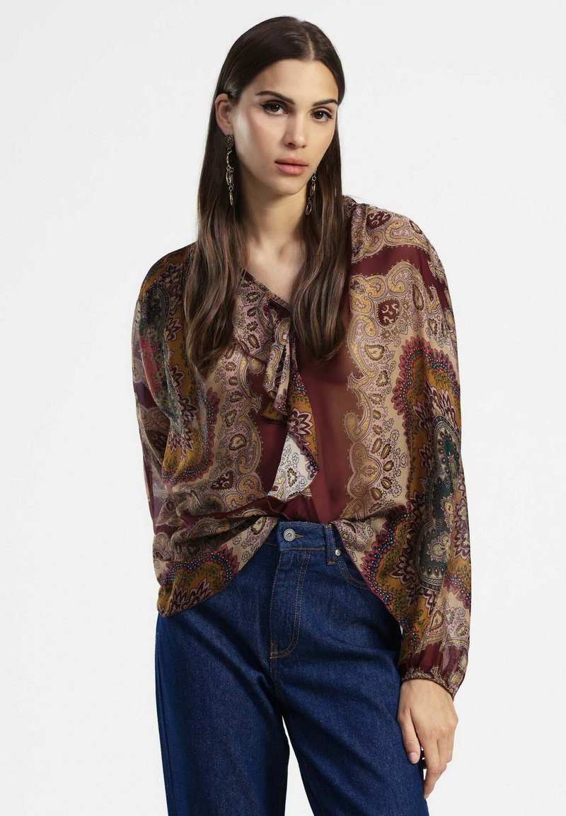 Blusa trasparente marrone con motivo paisley e scollatura legata; abbinata a jeans in denim a vita alta blu e orecchini pendenti.