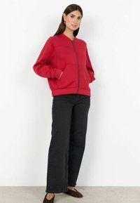 Felpa rossa con cerniera, scollo rotondo, polsini a coste e tasche a marsupio, abbinata a jeans neri a gamba larga e ballerine marroni.