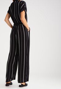 Černý pruhovaný overal má krátké rukávy, volný střih, široké nohavice a stáhnutý pas; látka je hladká a lehká.
