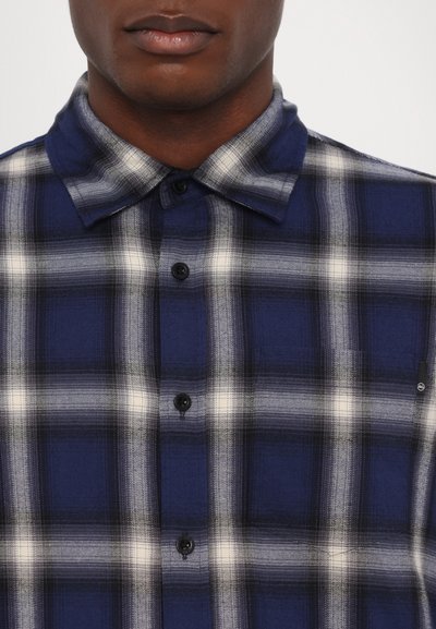 Tumedat sinine ja valge ruuduline särk nööpide all oleva kraega, mustade nööpidega ja sileda tekstuuriga. Vasakul küljel on väike logo.