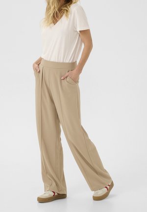 Kvinde iført beige vidde bukser, hvid kortærmet top og hvide sneakers med røde detaljer, står med hænderne i lommerne.