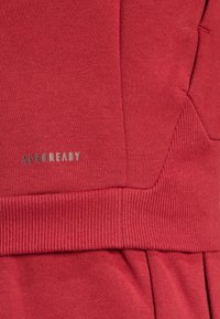 Red athletic garment featuring a soft, textured fabric with ribbed cuffs. "AEROREADY" branding in silver located on the front.