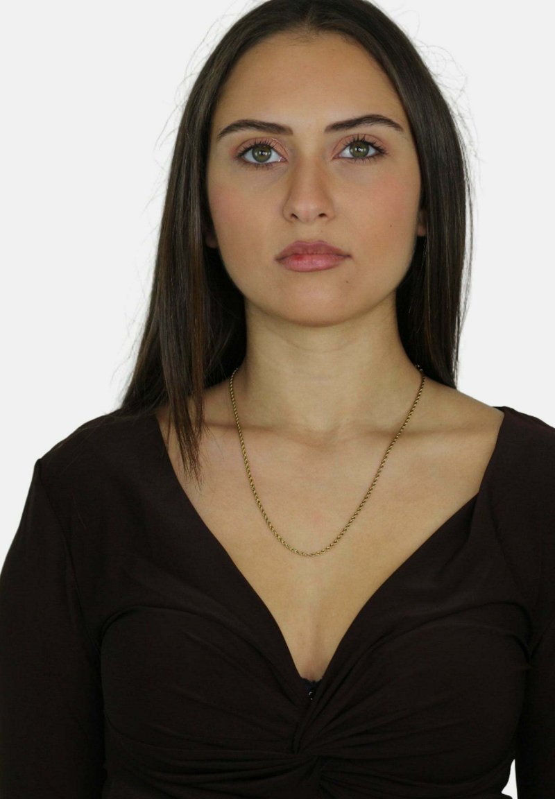 Giovane donna con capelli scuri e lisci, che indossa una maglietta nera a scollatura a V e una sottile collana d'oro, rivolta in avanti con un'espressione neutra.