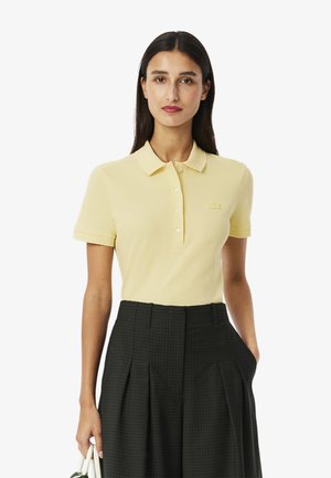Geel polo shirt met een klassieke kraag, vijf knopen en een klein geborduurd logo. Gebreide stof met een gladde textuur. G draagt het met geplooide donkere broeken.